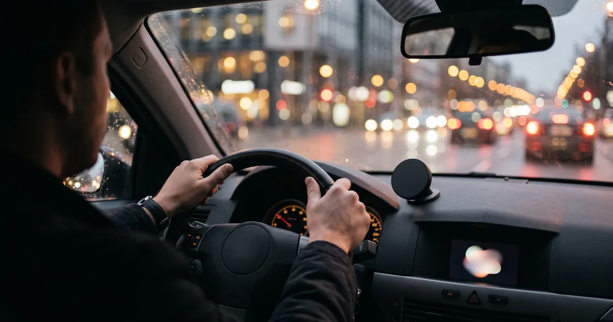 A car interior at dusk on a city street, the driver's hands on the steering wheel, a small plain round device mounted on the dashboard, city lights as bokeh thr