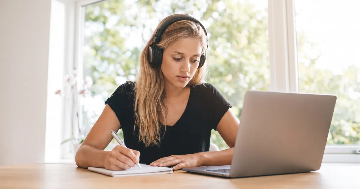 A young woman with long blonde hair and wireless over-ear headphones studying at a wooden desk by a bright window, writing in a notebook with an open laptop in front of her.