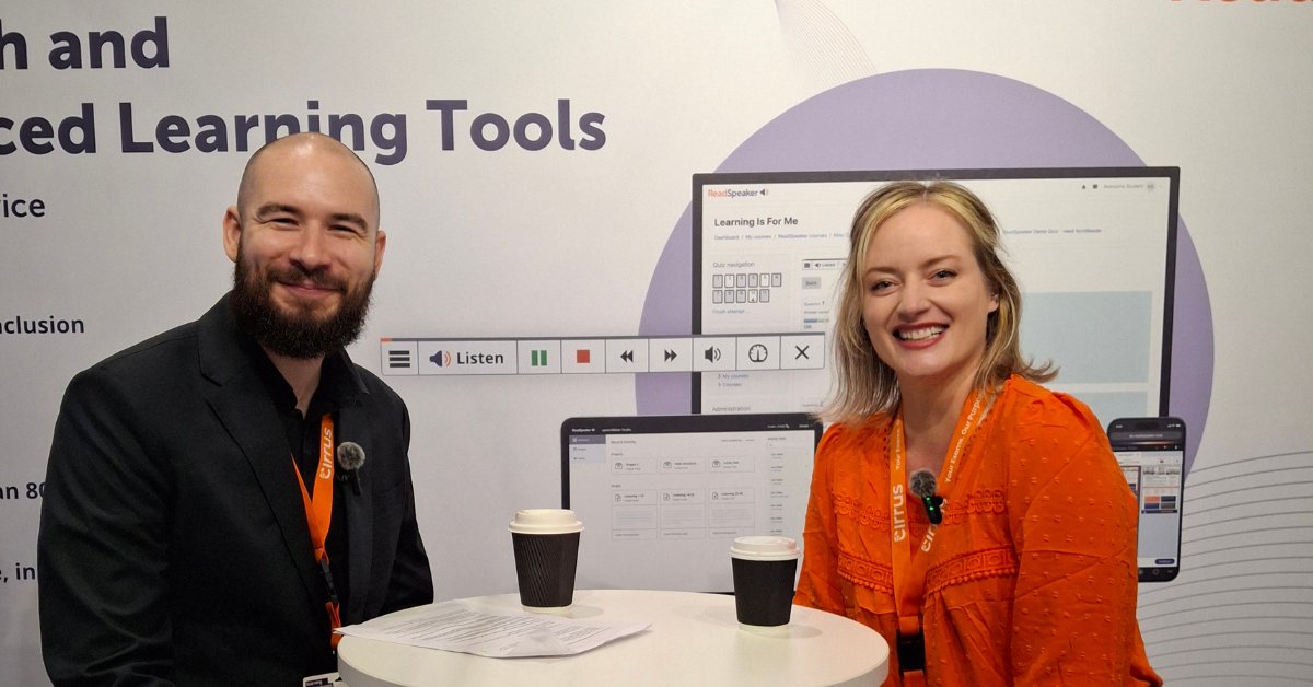 An image of two people, a man and a woman, sitting at a white pedestal table in front of a backdrop. The backdrop features text that includes "Learning Tools" and the name "Cirrus". The man is on the left, wearing a black blazer and an orange lanyard. The woman is on the right, wearing an orange top and a matching orange lanyard. Two coffee cups and some papers are on the table between them. In the background, there is a large screen displaying a digital interface with the text "Learning For Me" and a ReadSpeaker "Listen" button.