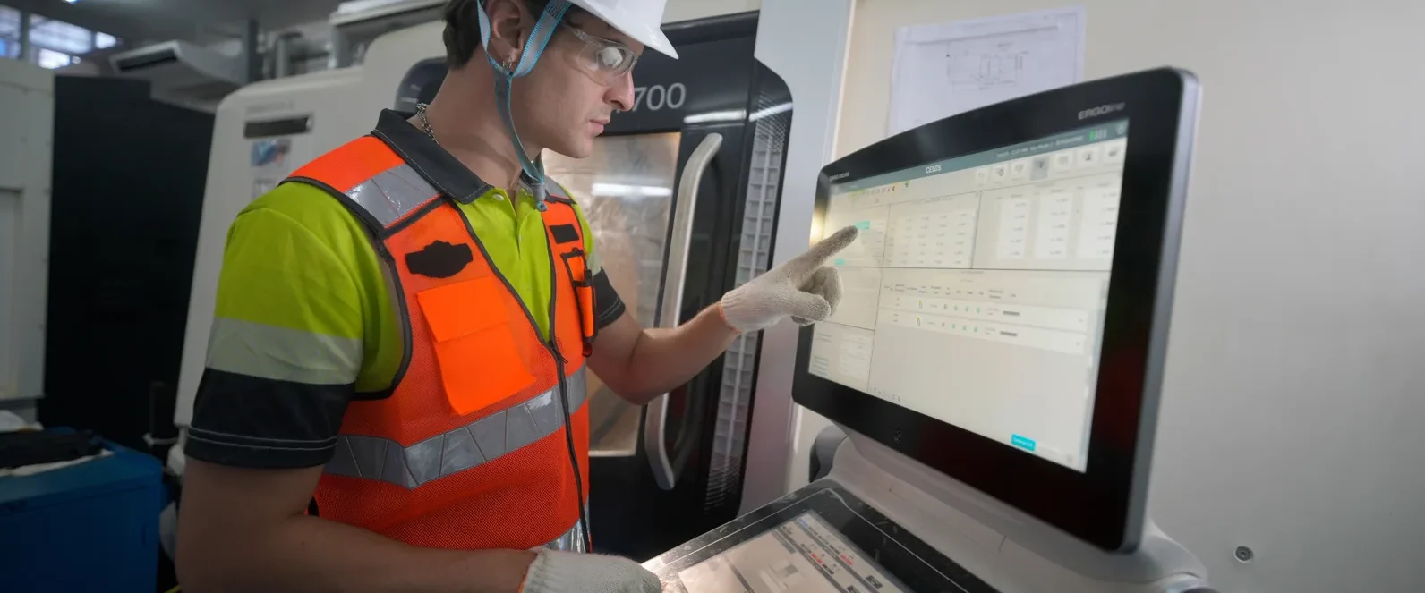 Technician in a hard hat and safety vest touching the screen of an industrial machine operator panel.