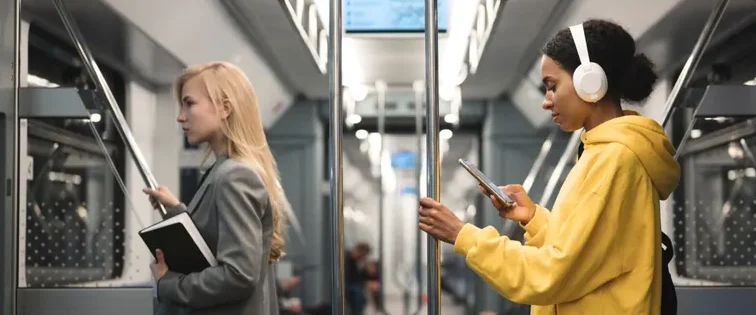 Two women in a subway, one reading and the other listening to music on headphones.
