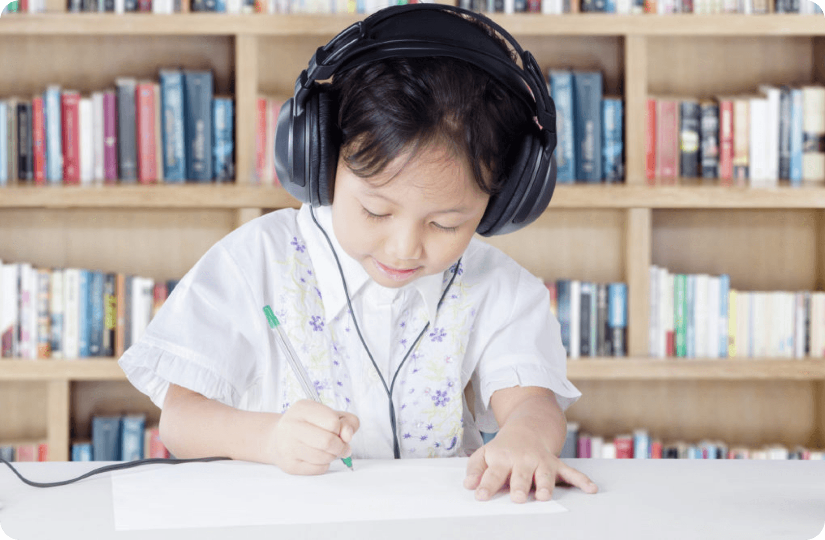 Criança usando fones de ouvido, desenhando no papel em uma biblioteca com estantes de livros ao fundo.