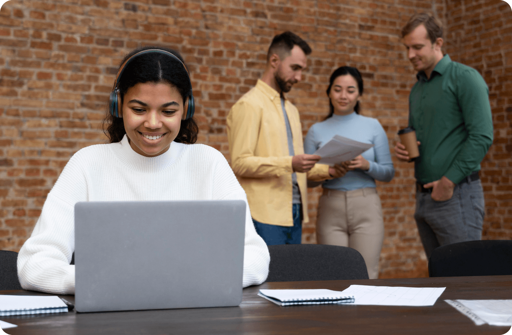 Smiling woman in headphones working on a laptop with colleagues discussing documents behind her.