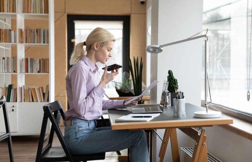 Una donna seduta a una scrivania con sopra un laptop utilizza uno smartphone.