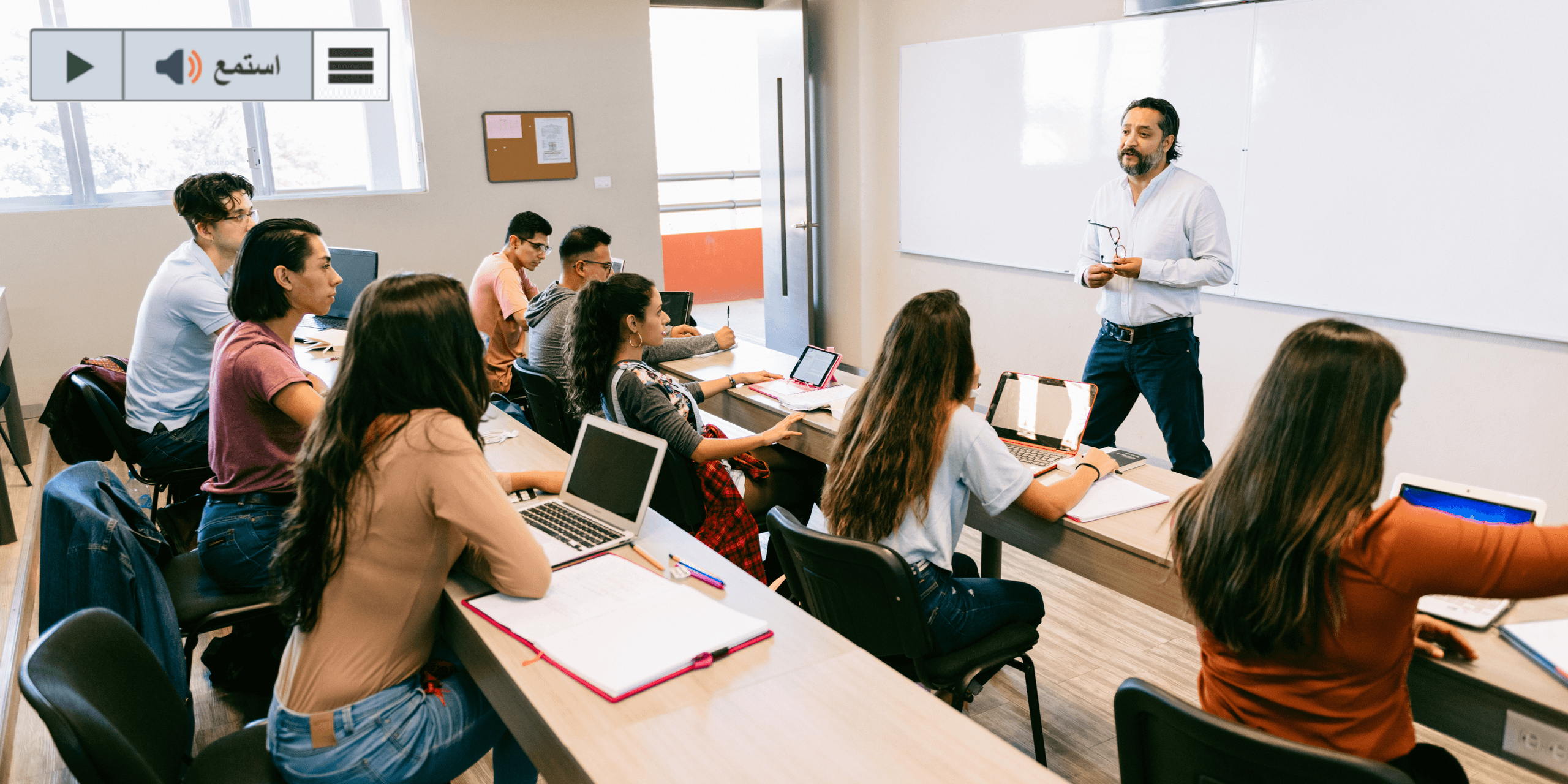 Students in a classroom listen to the teacher standing at the board.