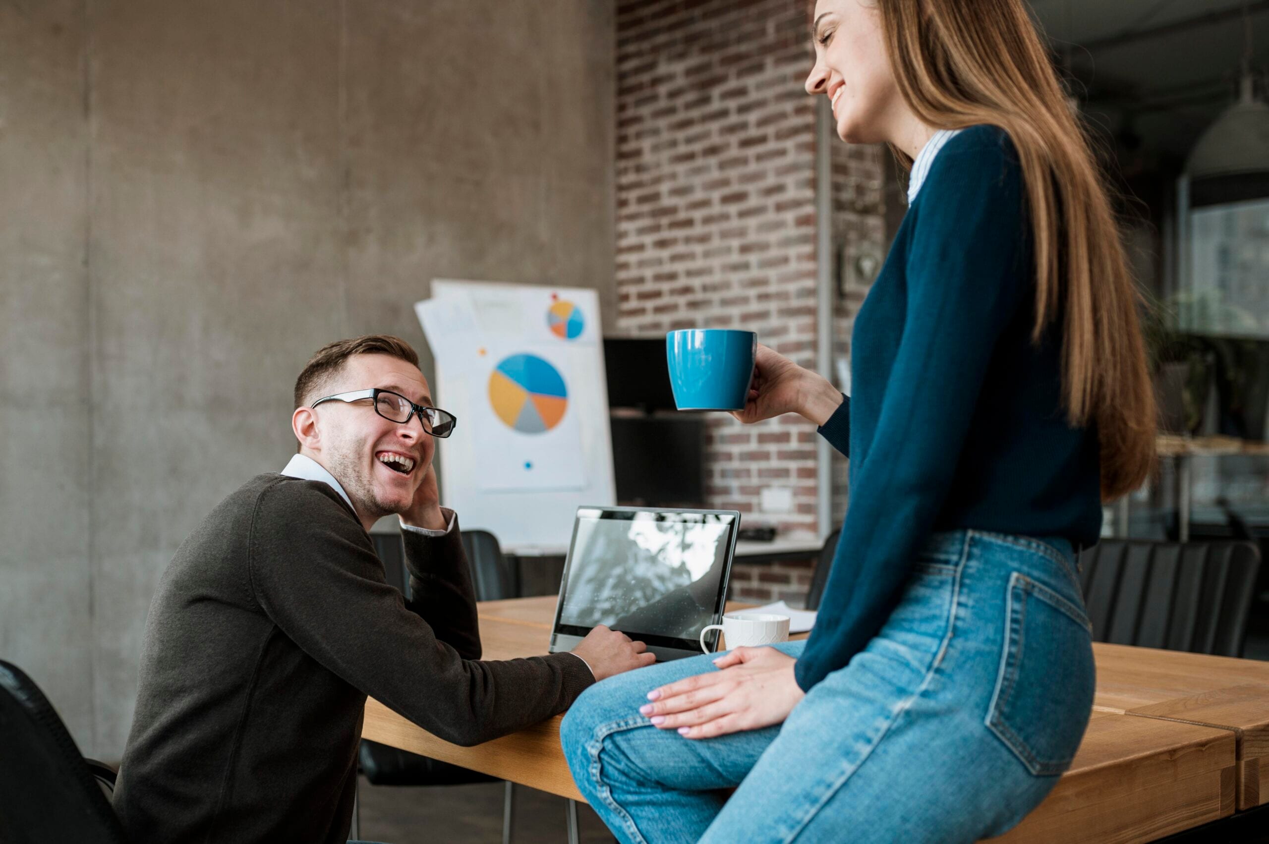 Two colleagues having a friendly conversation in a modern office setting; a man is seated at a table with a laptop, smiling, while a woman sits on the table holding a blue coffee mug. A presentation with pie charts is visible in the background.