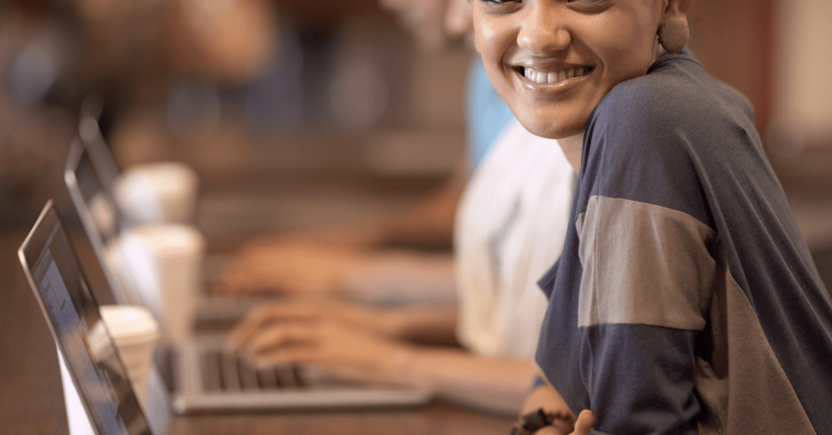 Smiling learner sitting at a laptop, with others typing on computers in the background.