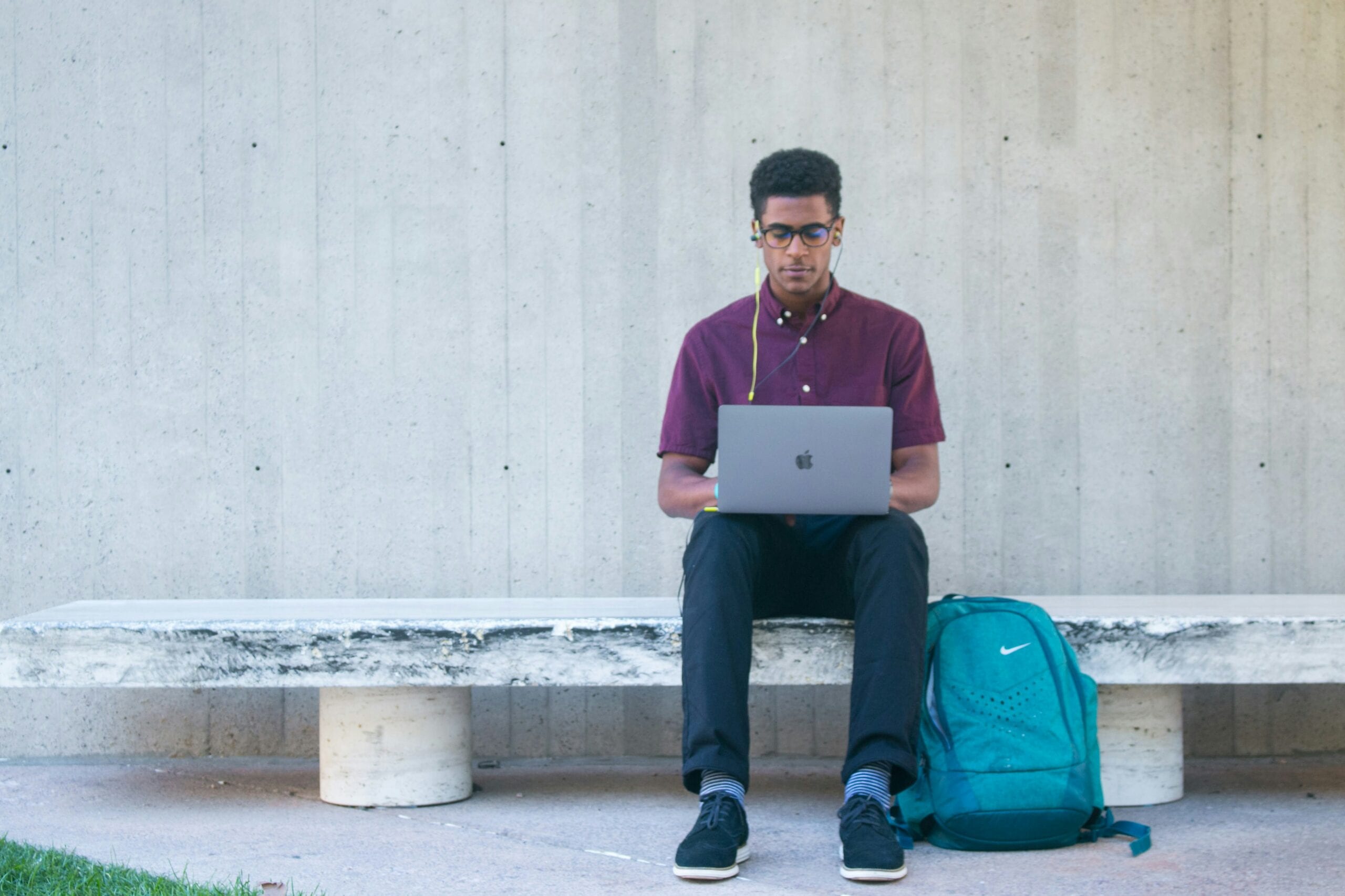 A man sitting on a bench with a laptop and blue backpack, wearing glasses and a purple shirt.