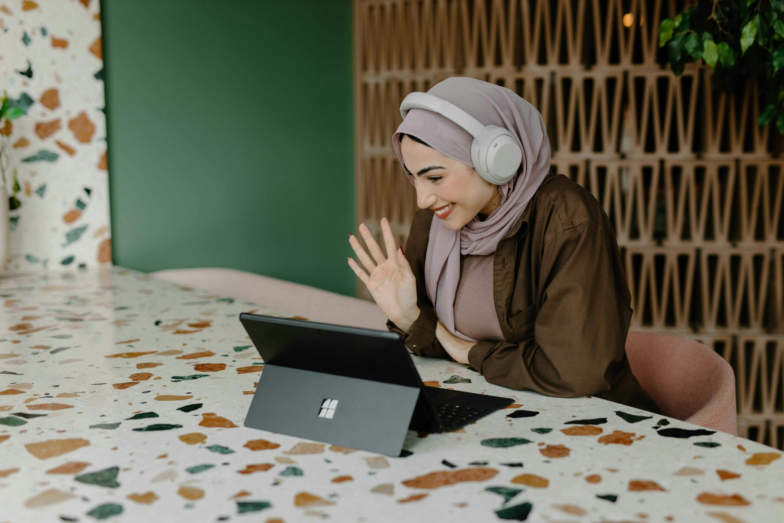 A woman wearing white headphones and a hijab, sitting at a table and waving her hand while watching a laptop.