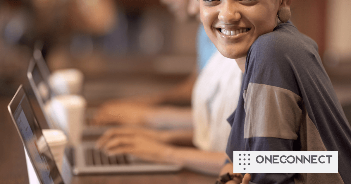 A woman smiles at the camera while typing on a laptop in a cafe. Warm ambient lighting and blurred background suggest a relaxed atmosphere. The text "ONECONNECT" is in the corner.
