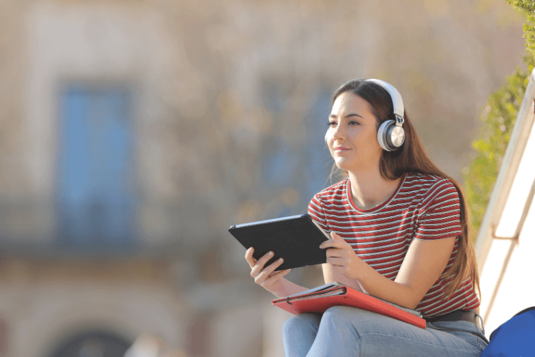 Girl with tablet wearing headphones sitting on a college campus building's stairs.