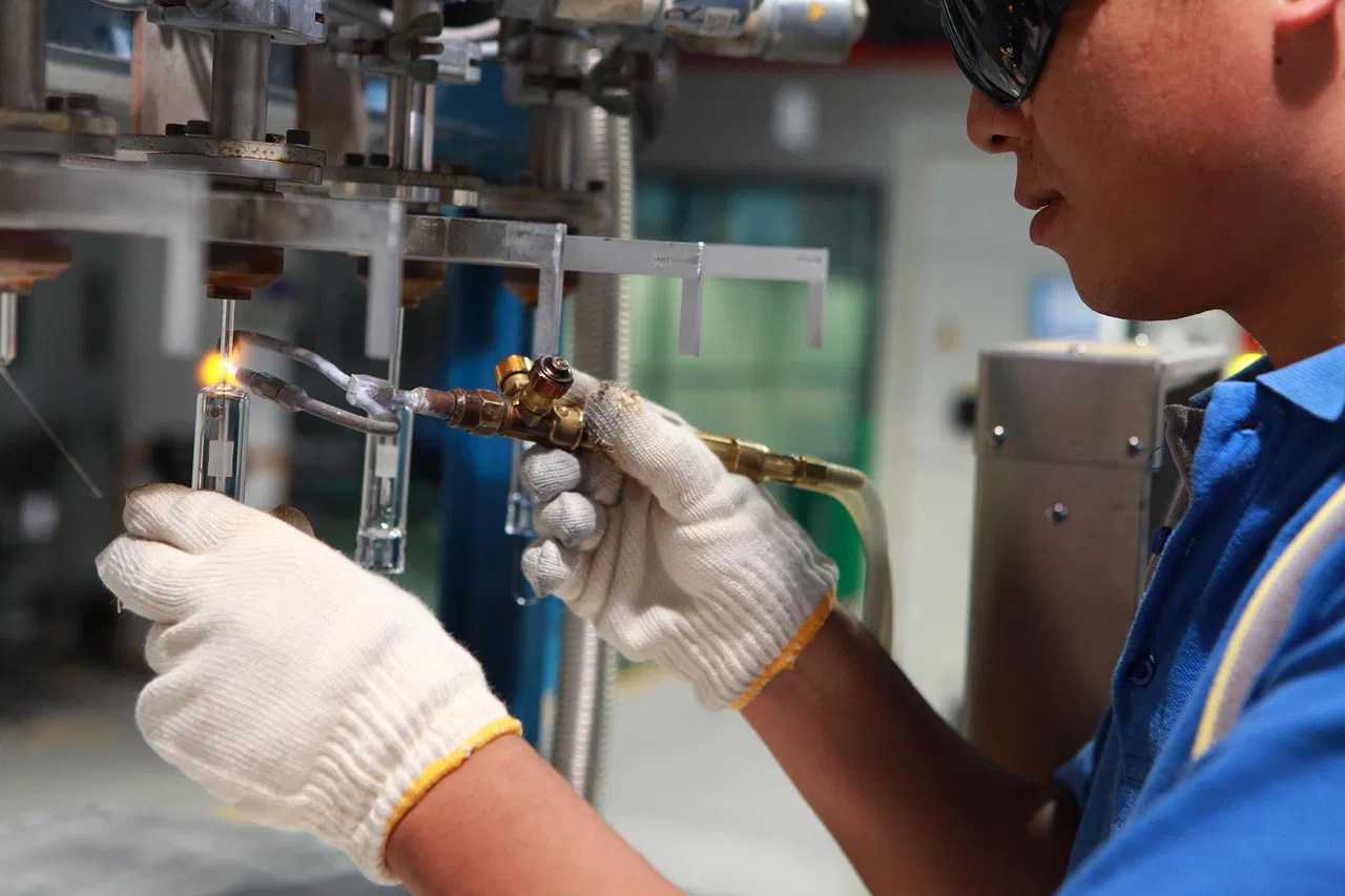 Un travailleur avec des gants et des lunettes de sécurité utilisant une torche à souder pour assembler des éléments en verre dans un atelier industriel.