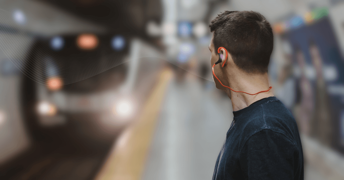 A wide horizontal banner featuring a man from the side, seen from the chest up, standing on a blurred subway platform as a train approaches in the background. He is wearing a dark t-shirt and bright orange wired earbuds. A subtle, white flowing wave sound line pattern emanates from his ear conveying a transport announcement, moving toward the left side of the frame across the subway station setting.