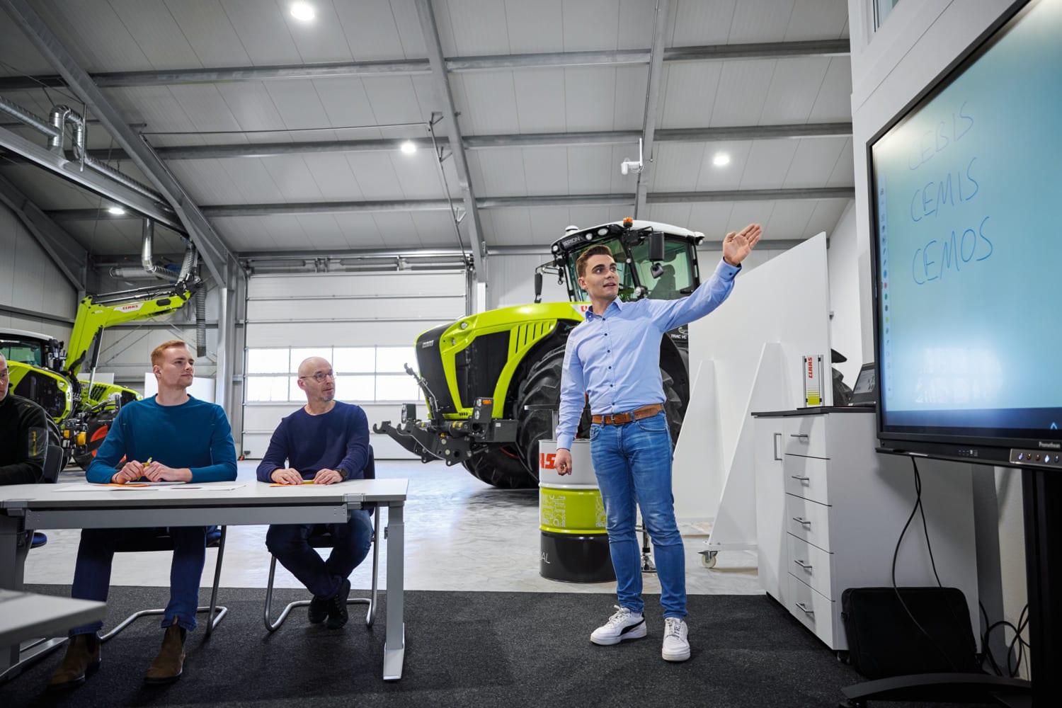 A trainer presenting in a CLAAS workshop, a tractor behind him