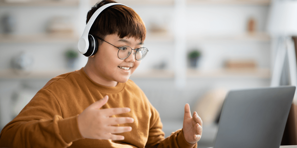 A boy of around 12 or 13 sits looking and smiling at a laptop. He is wearing headphones and his hands are gesturing towards the screen.
