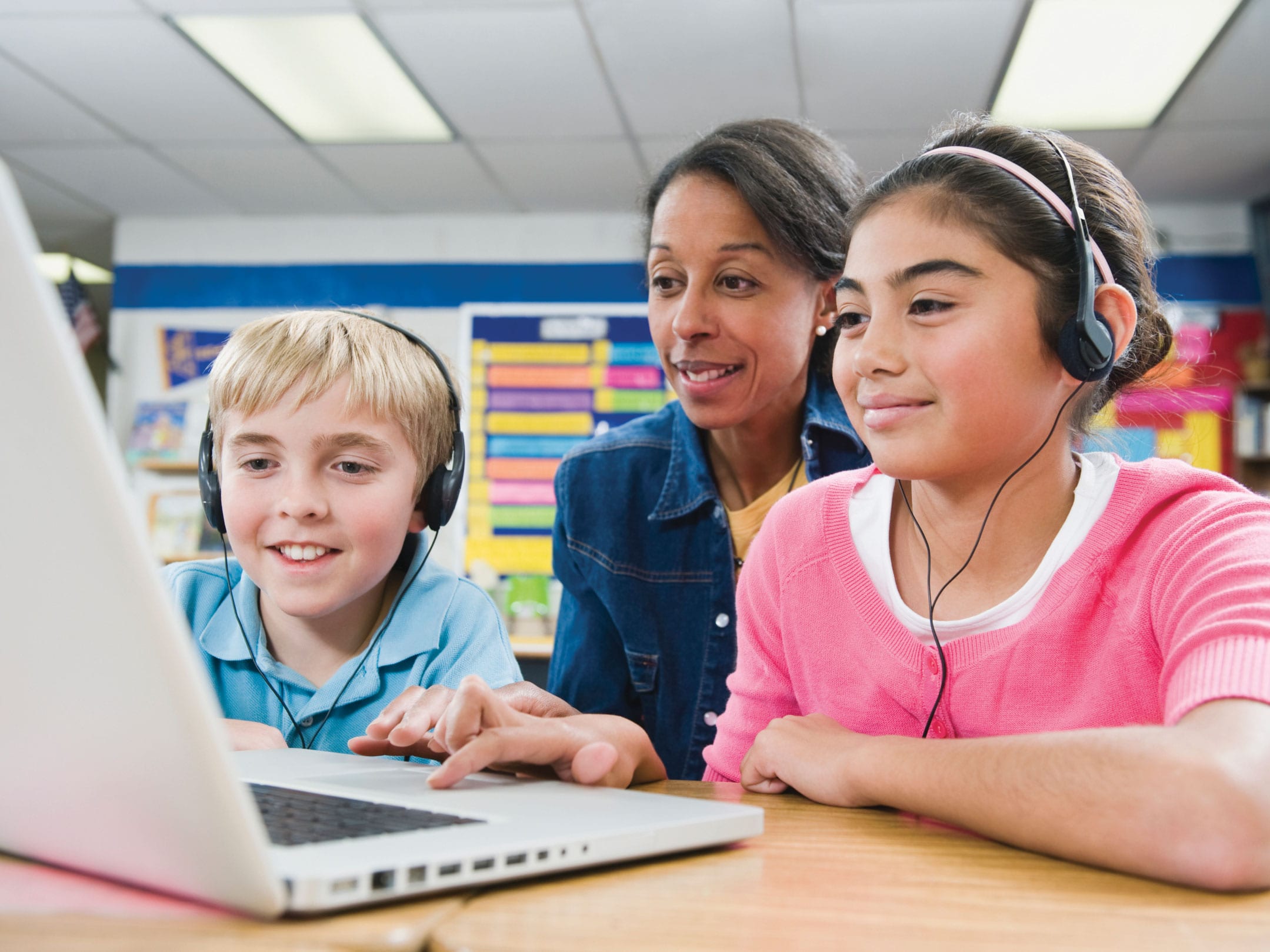A teacher and two pupils wearing headphones working together on a laptop in a classroom.